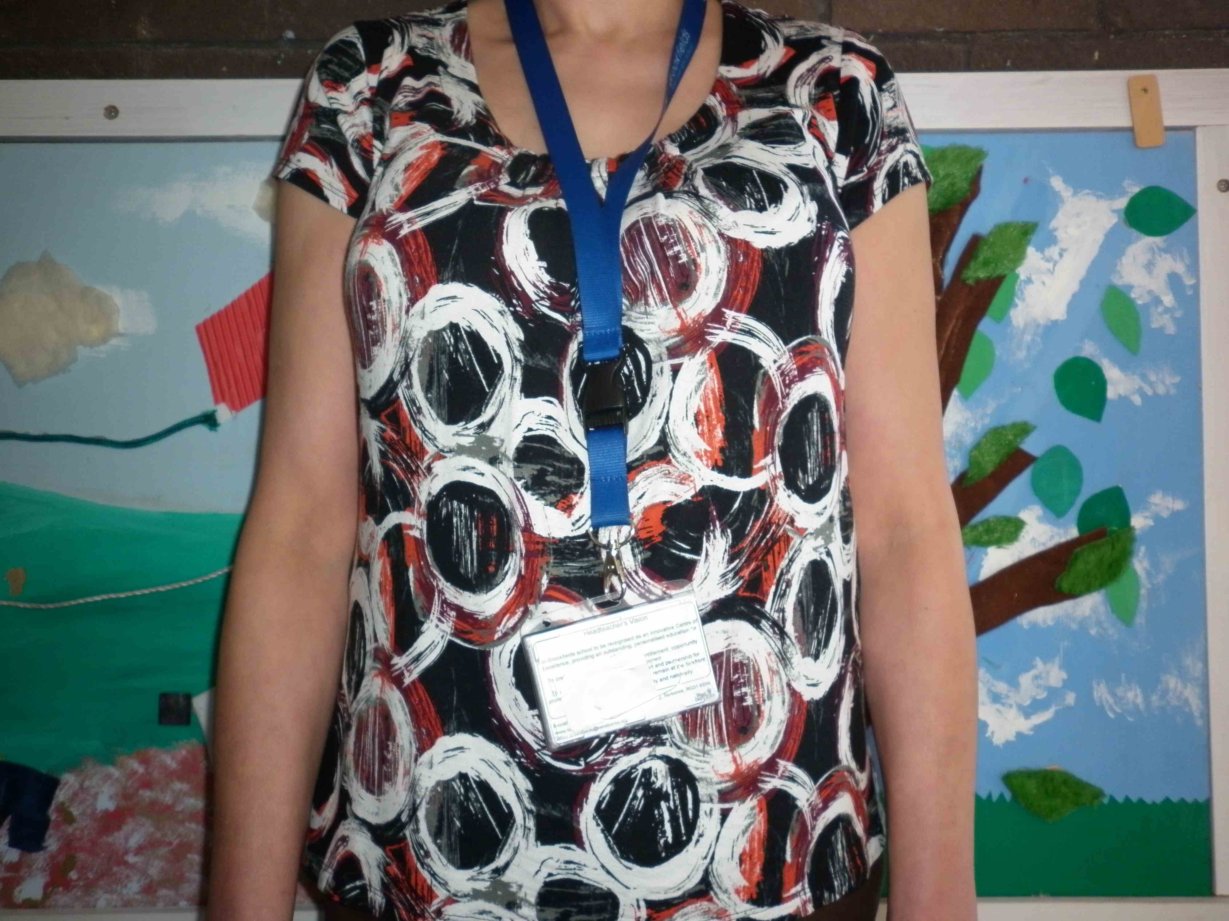 A woman's trunk. She is wearing a highly patterned dress, which provides poor visual contrast with her hands. Her ID badge is showing. She is standing in front of a visually distracting background.